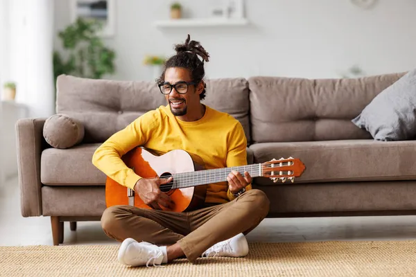 Genç Afro-Amerikalı adam evde akustik gitar çalıyor, oturma odasında yerde oturuyor.