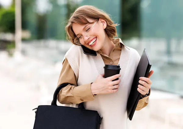 Jovem sorridente fêmea fica do lado de fora no parque da cidade com café enquanto fala no celular — Fotografia de Stock