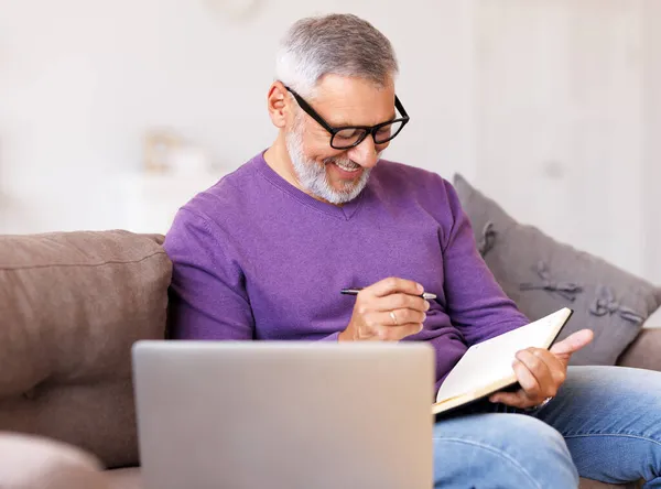 Guapo feliz hombre mayor en gafas de trabajo a distancia mientras está sentado en el sofá con el ordenador portátil —  Fotos de Stock