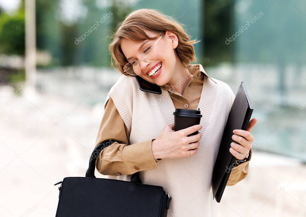 Young smiling female stands outside in city park with coffee while speaking on mobile phone