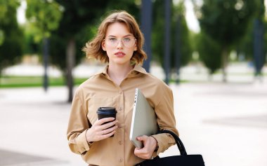 Kendine güvenen, iş kıyafeti giymiş kadın ofis çalışanı dışarıda dizüstü bilgisayar ve kahveyle bekliyor.