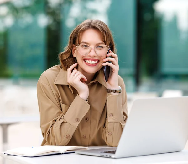 Office employee talking on cell phone with smile sitting on terrace in cafe with coffee and laptop — Stock Photo, Image