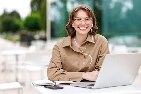 Glad ung kvinnlig lärare i hörlurar sitter framför laptop utanför har lektion på nätet — Stockfoto