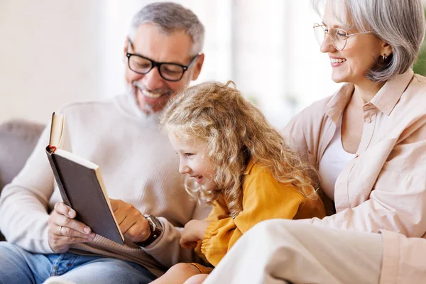 Cute little girl granddaughter smiling when reading book with senior grandparents — Stock Photo, Image