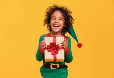 Aşırı neşeli Afrikalı Amerikalı çocuk Noel elf kostümü giyiyor ve elinde Noel hediyesi var.