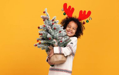 Kırmızı geyik boynuzları takmış küçük Afrikalı Amerikalı çocuk elinde karlı Noel ağacı tutuyor.