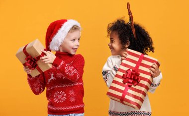 Noel kıyafetli mutlu çoklu etnik çocuklar ellerinde sarı arka planda Noel hediyeleri tutuyorlar.