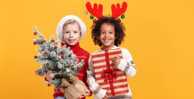 Mutlu çok ırklı çocuklar Noel Baba şapkası ve geyik boynuzları takıp ellerinde karlı Noel ağacı ve hediye kutusu tutuyorlar.