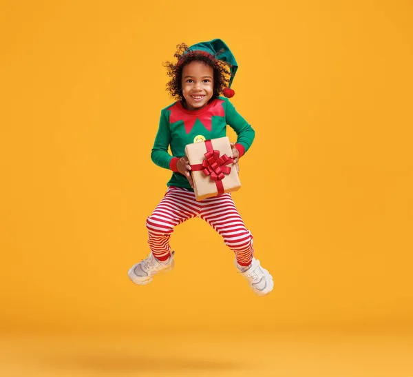 Feliz afroamericano niño traje de elfo de Navidad con regalo de Navidad envuelto en manos saltando en el aire — Foto de Stock