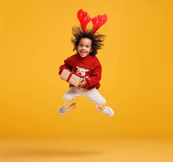 Emocionado niño afroamericano en jersey de Navidad y cuernos de ciervo saltando en el aire con la caja de regalo de Navidad — Foto de Stock