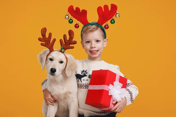 Ευτυχισμένο παιδί προσχολικής ηλικίας με Χριστουγεννιάτικο δώρο αγκαλιάζει το κουτάβι Golden Retriever, φορώντας κόκκινα κέρατα ταράνδου — Φωτογραφία Αρχείου