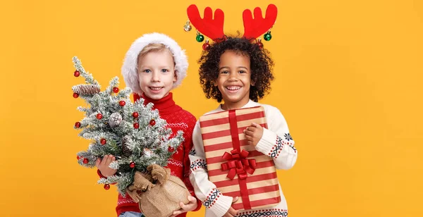 Crianças multirraciais felizes usando chapéu de Papai Noel e chifres de veado segurando árvore de Natal nevado e caixa de presente — Fotografia de Stock