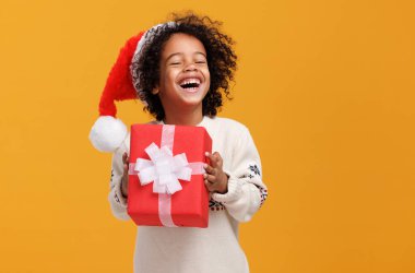 Noel Baba şapkası ve örgü süveter ören kıvırcık saçlı mutlu küçük Afro-Amerikan çocuk büyük Noel hediye kutusunu sıkıca sarılıyor.