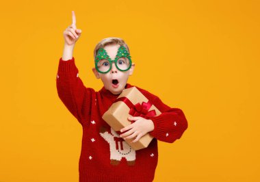 Noel ağacı şeklinde komik gözlük takan gülümseyen küçük çocuk Noel hediyesini parmağıyla yukarı kaldırarak gösteriyor.