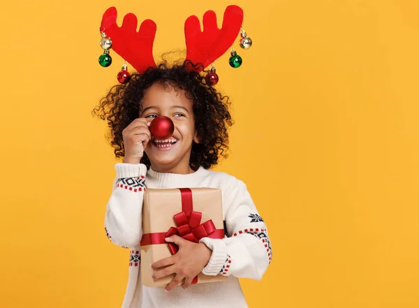 Χαρούμενο μικρό εθνοτικό αγόρι με τυλιγμένο χριστουγεννιάτικο δώρο κρατώντας κόκκινη μπάλα χριστουγεννιάτικο δέντρο μπροστά από τη μύτη του — Φωτογραφία Αρχείου