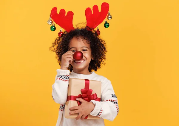 Alegre niño étnico con regalo de Navidad envuelto sosteniendo bola de árbol de Navidad rojo en frente de su nariz — Foto de Stock