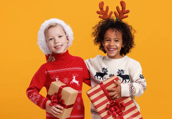 Χαρούμενα ενθουσιασμένα πολυφυλετικά παιδιά με Χριστουγεννιάτικο ντύσιμο κρατώντας χριστουγεννιάτικα δώρα σε κίτρινο φόντο — Φωτογραφία Αρχείου