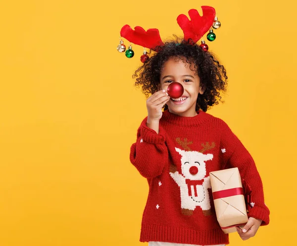 Alegre niño étnico con regalo de Navidad envuelto sosteniendo bola de árbol de Navidad rojo en frente de su nariz — Foto de Stock