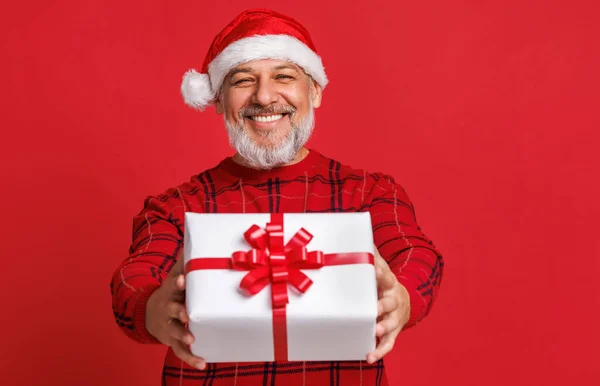 Kırmızı Noel Baba şapkalı neşeli yaşlı adam elinde bir Noel hediyesi tutuyor ve renkli kırmızı bir arka plan ile gülüyor. — Stok fotoğraf