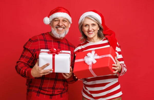 Kırmızı Noel Baba şapkası takmış son sınıf aile çifti ellerinde bir Noel hediyesi tutuyor ve renkli kırmızı bir arka plan ile gülüyorlar. — Stok fotoğraf