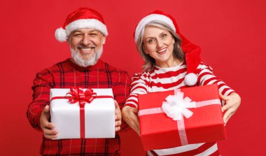  Kırmızı Noel Baba şapkası takmış son sınıf aile çifti ellerinde bir Noel hediyesi tutuyor ve renkli kırmızı bir arka plan ile gülüyorlar.