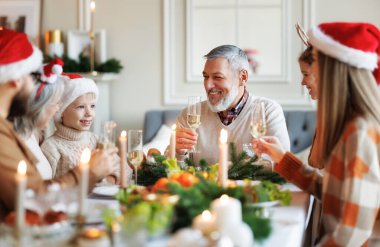 Noel Baba şapkalı mutlu bir aile yemek masasında kadeh kaldırıyor ve kadeh tokuşturuyor.