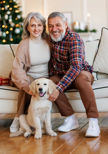 Gelukkig senior familie paar zitten in de buurt van versierde kerstboom thuis met hond golden retriever puppy — Stockfoto