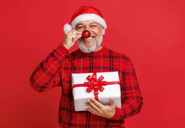 Šťastný legrační senior muž v Santa klobouk s dárkovou krabici drží červené vánoční strom míč před nosem — Stock fotografie