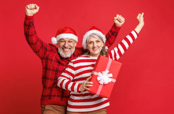 Happy euphoric senior retired family couple in Santa hats with wrapped Christmas gift raising hands — Stock Photo, Image
