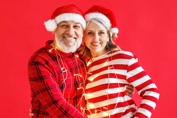 クリスマスライトと赤い背景のサンタの帽子の幸せなシニア家族のカップルは新年を祝う — ストック写真