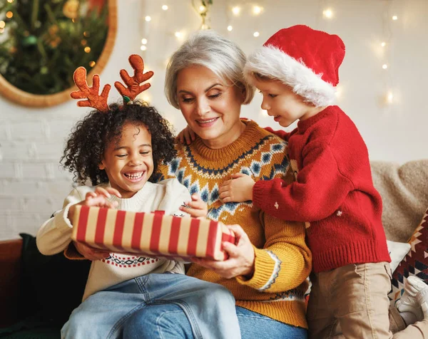 Zwei Jungen verschiedener Rassen überreichen der Großmutter ein Neujahrsgeschenk, während sie am Weihnachtsbaum sitzen — Stockfoto