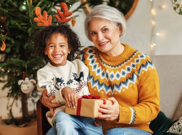 Glad afro amerikansk lockig pojke sitter på mormor knä med julklapp, ler mot kameran — Stockfoto