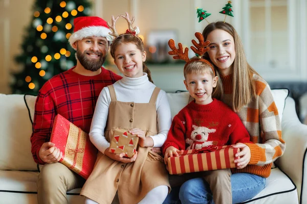 Jóvenes padres y niños felices con cajas de regalo sentados en el sofá cerca del árbol de Navidad decorado — Foto de Stock