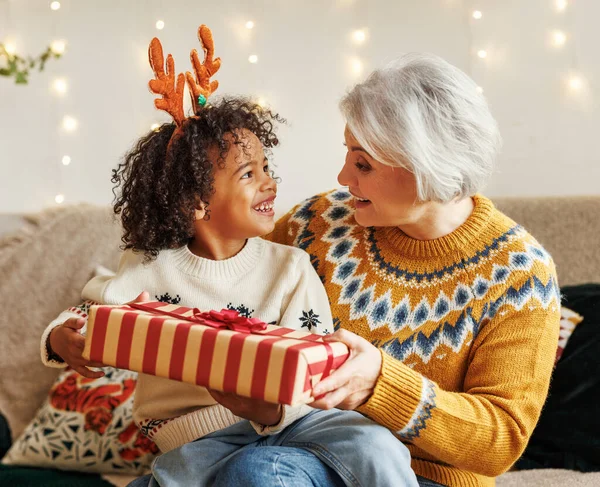 快乐的非洲卷曲男孩坐在祖母的膝上，带着圣诞礼物，在镜头前微笑 — 图库照片