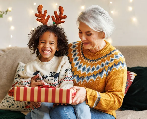 Glücklicher afroamerikanischer Lockenkopf sitzt mit Weihnachtsgeschenk auf Großmutters Schoß und lächelt in die Kamera — Stockfoto