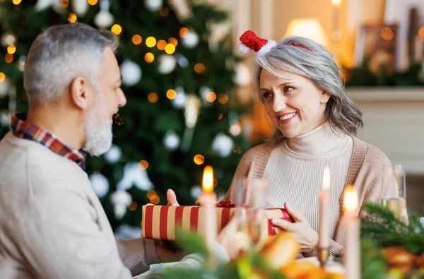 Happy senior family couple hugging and exchanging Christmas presents during festive dinner at home — Stock Photo, Image