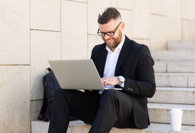 Genç mutlu iş adamı takım elbiseli, dışarıda merdivenlerde dizüstü bilgisayarı ve kahvesiyle oturuyor.