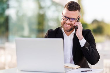 Gülümseyen işadamı cep telefonuyla keyifli sohbetler yapıyor, açık kafede kahve içiyor.