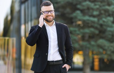  Gözlüklü mutlu iş adamı güneşli bir günde dışarıda yürüyor ve yakın arkadaşı ya da ailesi ile cep telefonundan konuşuyor. 