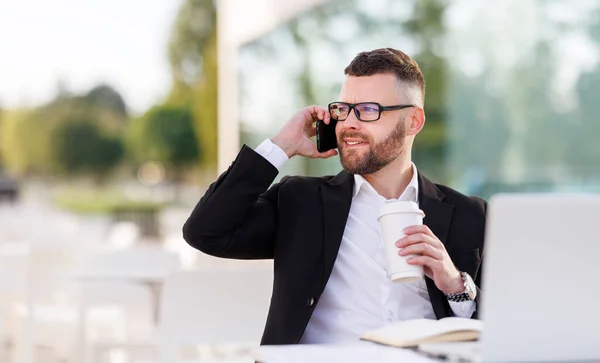 Glimlachende zakenman geniet van aangenaam gesprek op mobiele telefoon, het drinken van koffie in outdoor cafe — Stockfoto