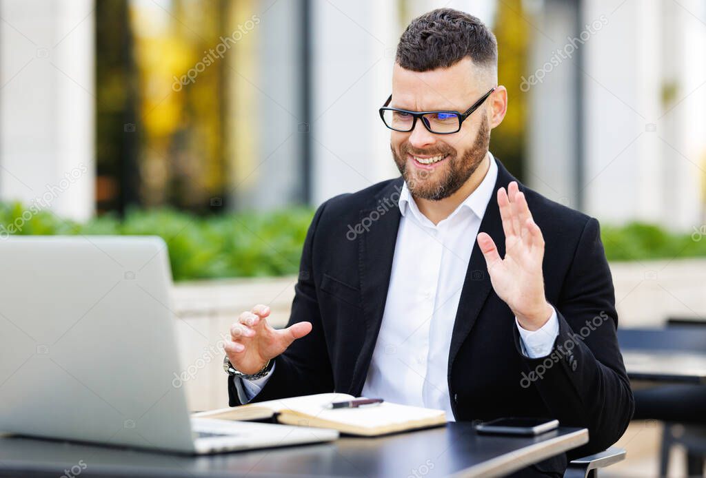 Happy young teacher in formal outfit and glasses giving lesson online sitting with laptop outside
