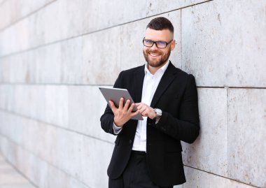 Takım elbiseli neşeli işadamı dışarıda beton duvarın önünde dikilirken dijital tablet kullanıyor.