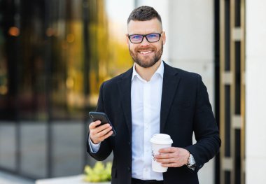 Resmi takım elbiseli gülümseyen genç ofis çalışanı dışarıda cep telefonuyla dikiliyor.