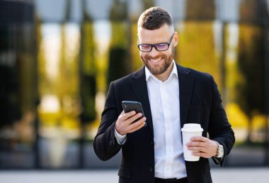 Resmi takım elbiseli gülümseyen genç ofis çalışanı dışarıda cep telefonuyla dikiliyor.