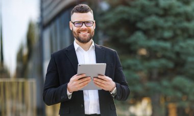 Neşeli iş adamı güneşli bir sokakta dikilirken dijital tablete bakıyor.