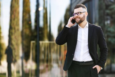 Gözlüklü genç işadamı güneşli bir günde dışarıda yürüyor ve yakın arkadaşı ya da ailesiyle cep telefonundan konuşuyor.