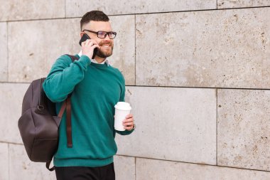 Genç, mutlu bir ofis çalışanı elinde kahveyle dışarıda cep telefonuyla konuşuyor.