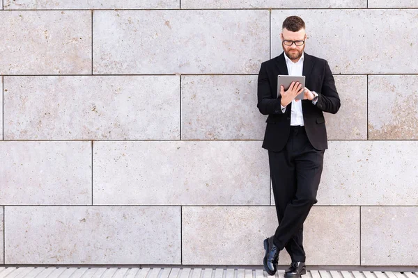 Hombre de negocios alegre en traje usando tableta digital mientras está parado afuera contra la pared de hormigón — Foto de Stock