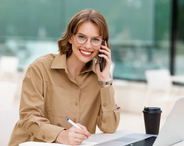 Feliz joven asistente de oficina trabajando remotamente en línea sentado con el ordenador portátil y café fuera — Foto de Stock