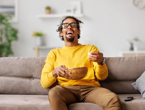 Fiatal túlzsúfolt afro-amerikai férfi popcornnal, aki hangosan nevet, és otthon vígjátékot néz. — Stock Fotó